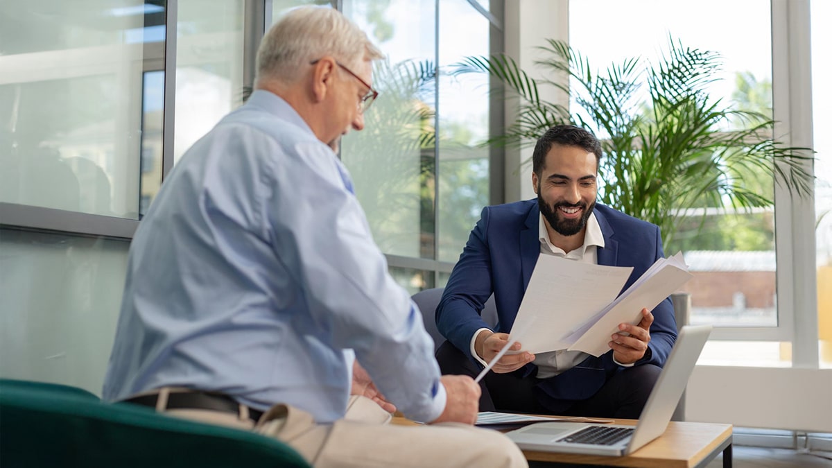 Business advisor smiling and reviewing documents with a senior client across a desk with a laptop